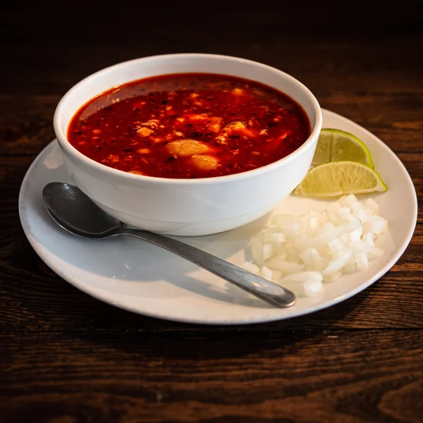A deep bowl of menudo in red chile broth, with diced onion and lime wedges on the plate beneath it.
