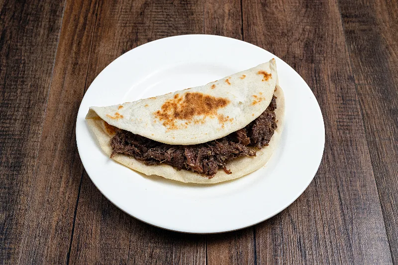A folded flour tortilla filled with barbacoa, on a white plate on a dark wood table.