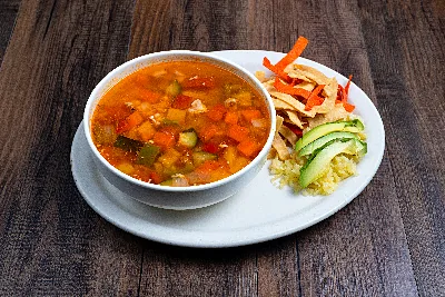 A bowl of tortilla soup with diced chicken and vegetables, served with tortilla strips, sliced avocado and cheese.