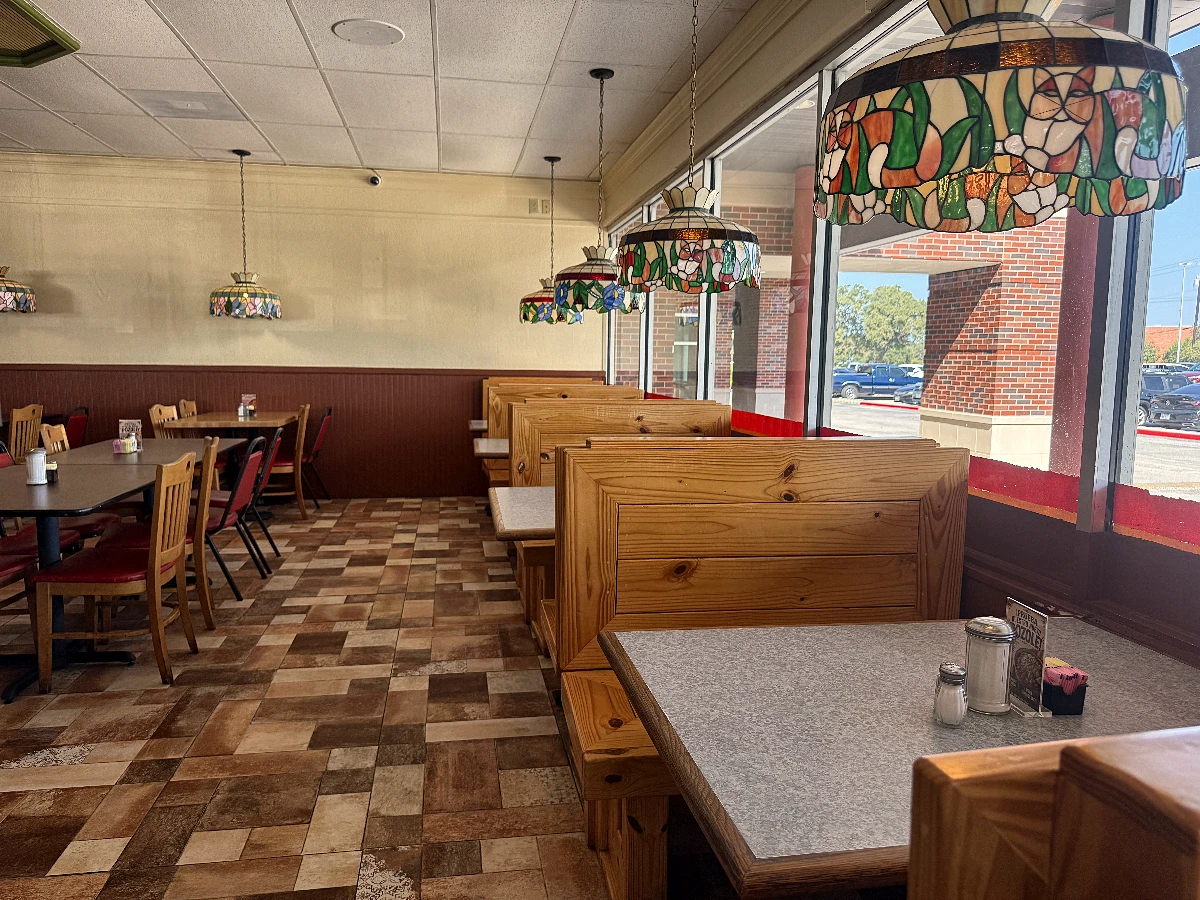 Pine booths facing the window wall beneath a line of stained-glass pendants, with the parking lot and a brick facade visible through the glass.